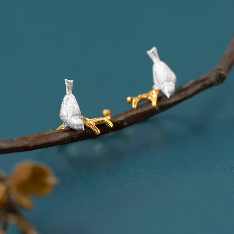 A pair of silver bird-shaped earrings with 18k gold plating, displayed on a branch with a blue background.