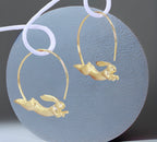 A pair of gold-plated hoop earrings with a silver rabbit design leaping out from the hoop.