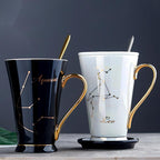 Two ceramic water cups, one black with gold accents and bone china handle, and one white with gold accents and a porcelain handle. Both cups have zodiac-related designs, with the black cup featuring Aquarius and the white cup featuring Leo.