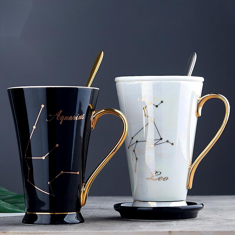 Two ceramic water cups, one black with gold accents and bone china handle, and one white with gold accents and a porcelain handle. Both cups have zodiac-related designs, with the black cup featuring Aquarius and the white cup featuring Leo.